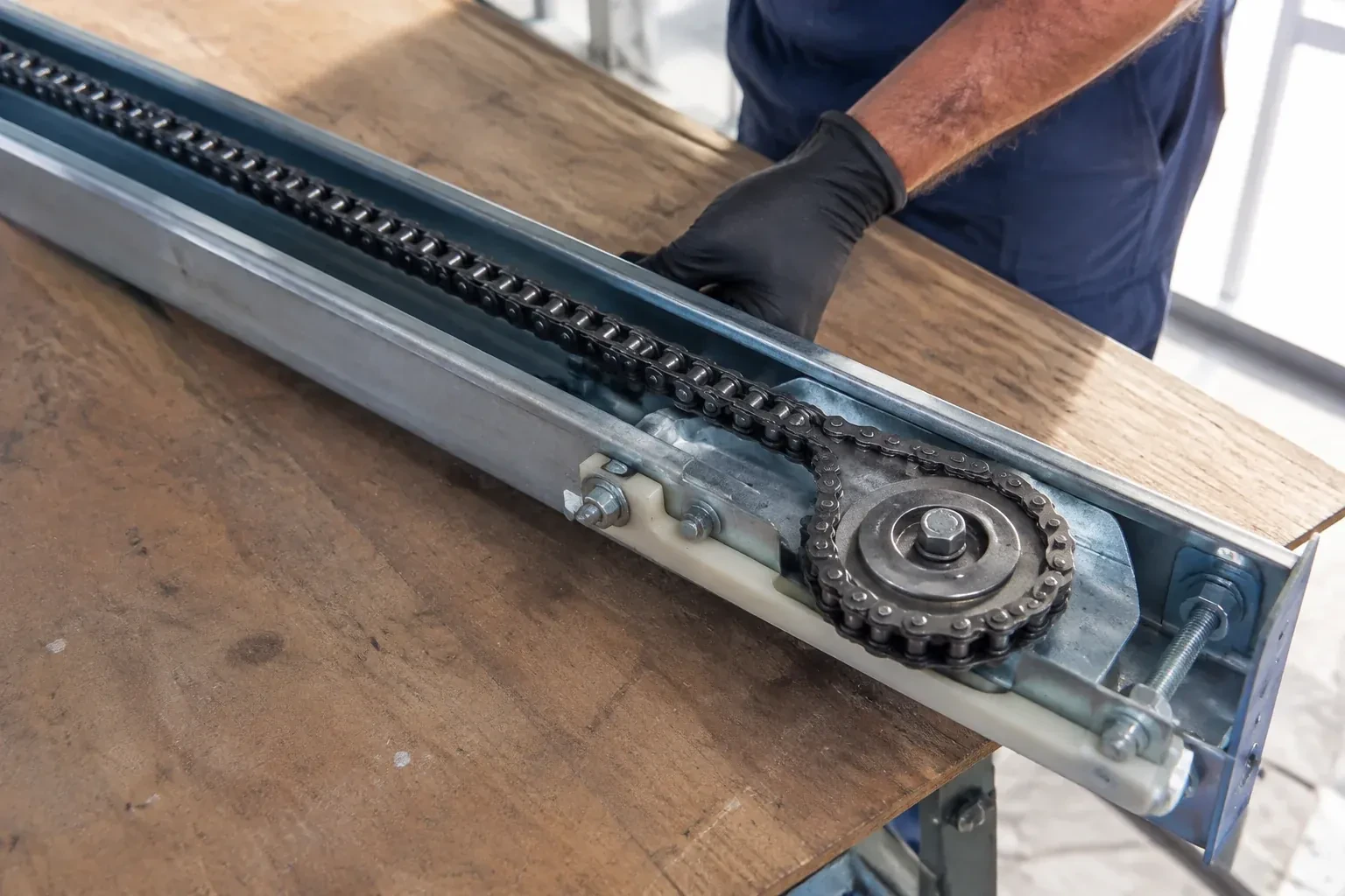 Gloved hand resting on a steel opener rail laid on a wooden bench, showing a drive chain looped around a sprocket wheel. — Chain-Drive Opener Repair, Centennial